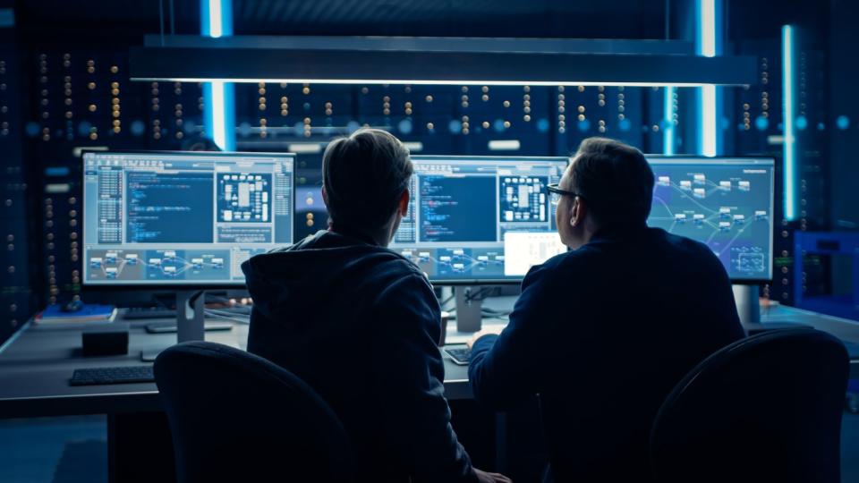 Two employees looking at copious amounts of data displayed on multiple computer monitors. 