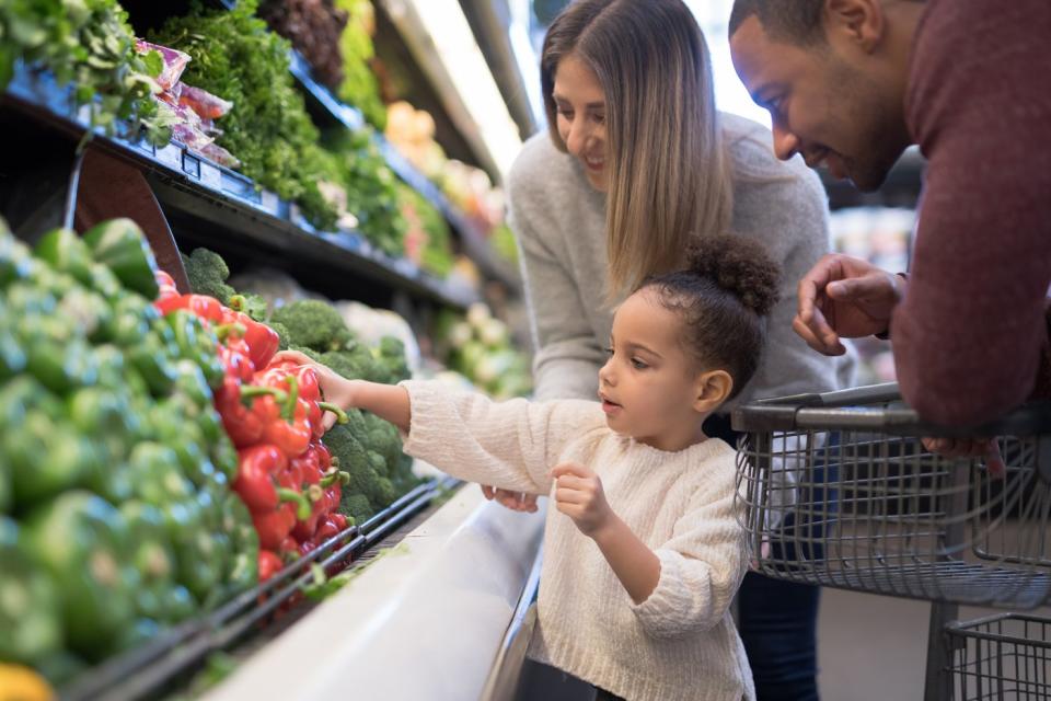 A child picking a bell pepper from the produce section of a grocery store, with parents standing nearby. 