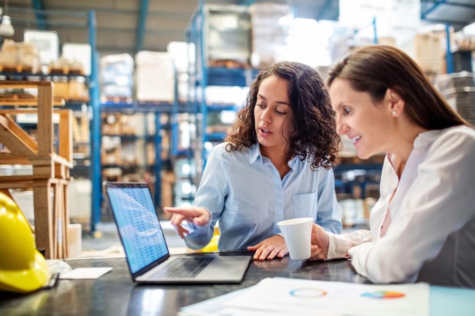 Two people in a warehouse sitting at a table discussing what is on a computer screen. 