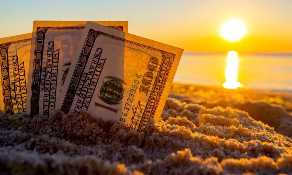 Three one hundred dollar bills stood up and partially buried in the sand, with the sun rising on the horizon.