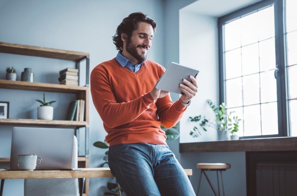 A person leans against a desk and looks at something on a tablet. 