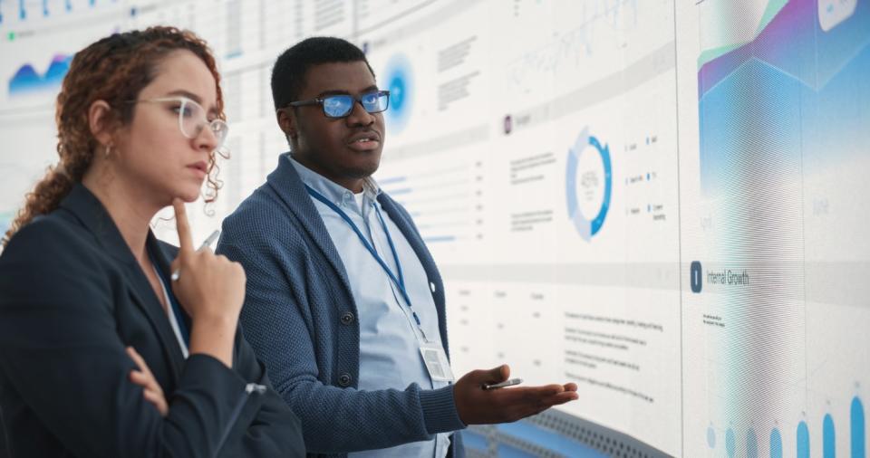 Two workers looking at data analytics on huge screen. 