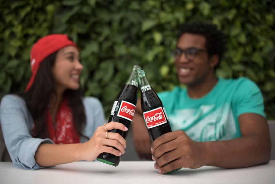 Two people clanking their Coca-Cola bottles together while seated outside and chatting.