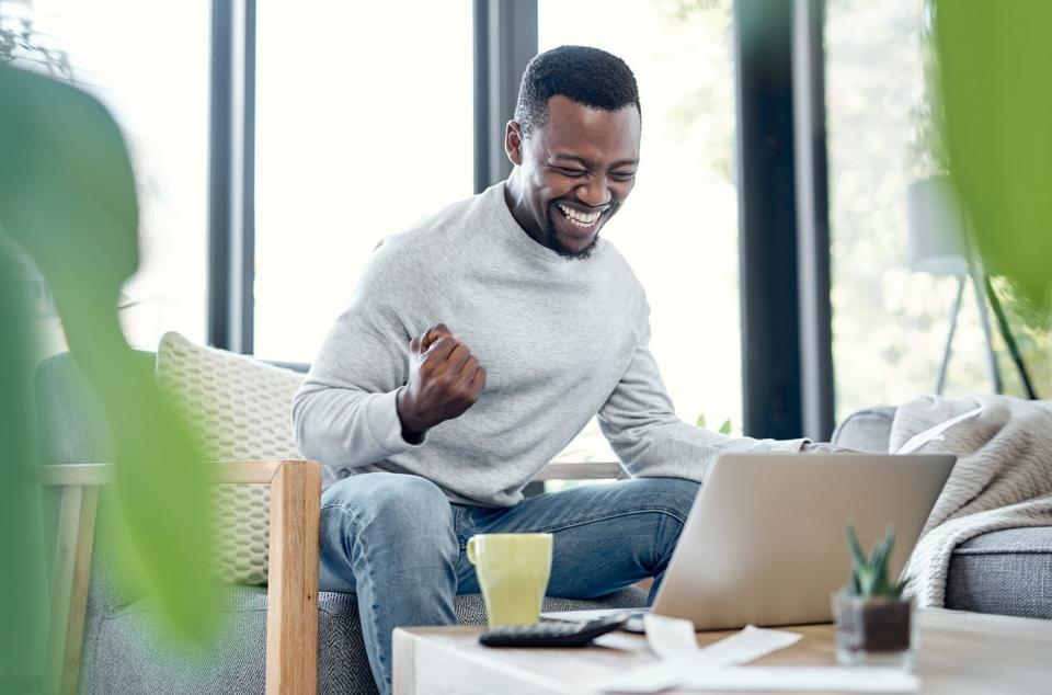 Someone is seated, looking at a laptop, and pumping his fist while smiling. 