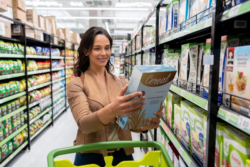 Shopper studies a box of cereal while walking through a brightly colored aisle in a grocery store.