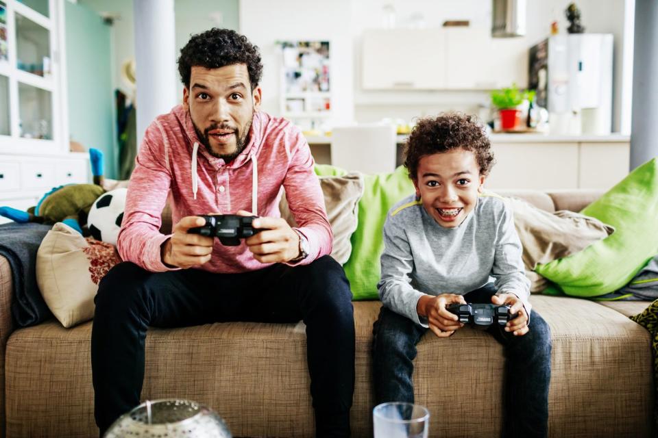 A parent and child seated on a couch and holding controllers while playing video games. 