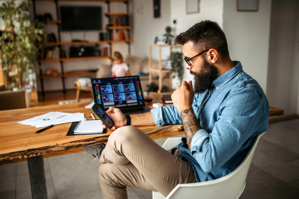 A person looking at stock charts on their smartphone with a laptop sitting on a table in the background.