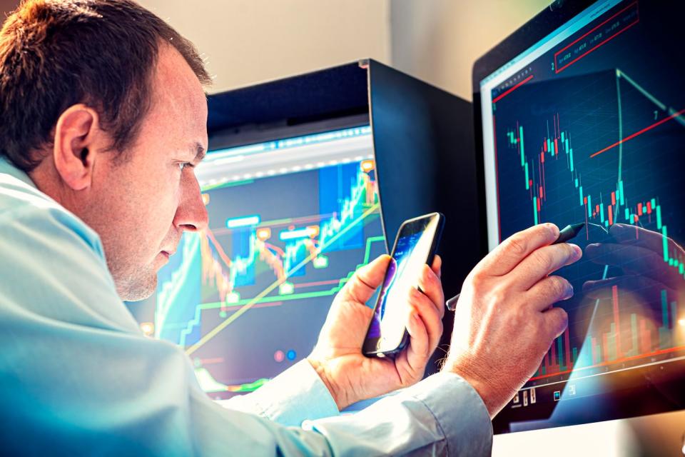 A money manager using a stylus and smartphone to analyze a stock chart displayed on a computer monitor. 